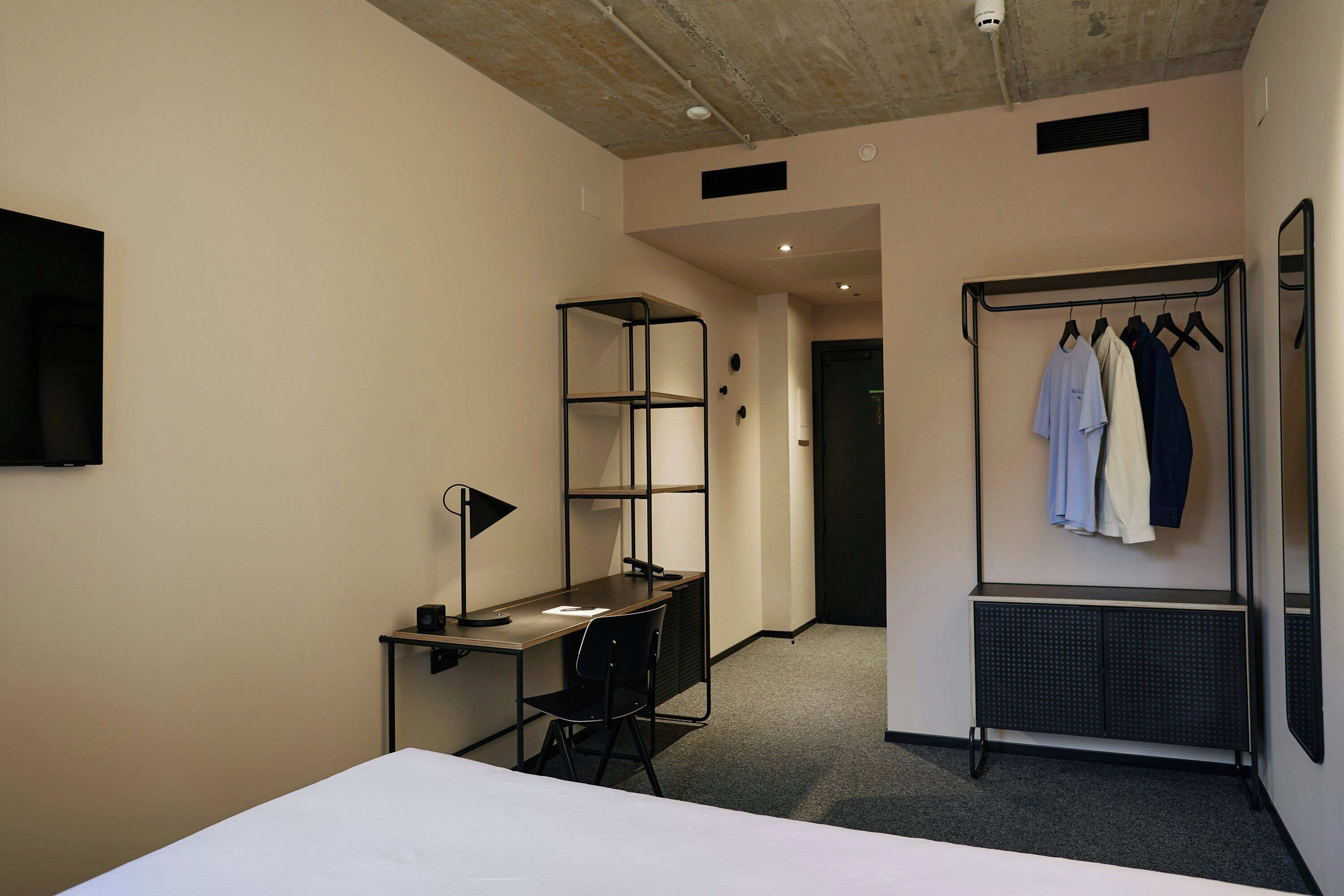 A hotel room with a bed, desk, chair, and a clothing rack with hangers.
