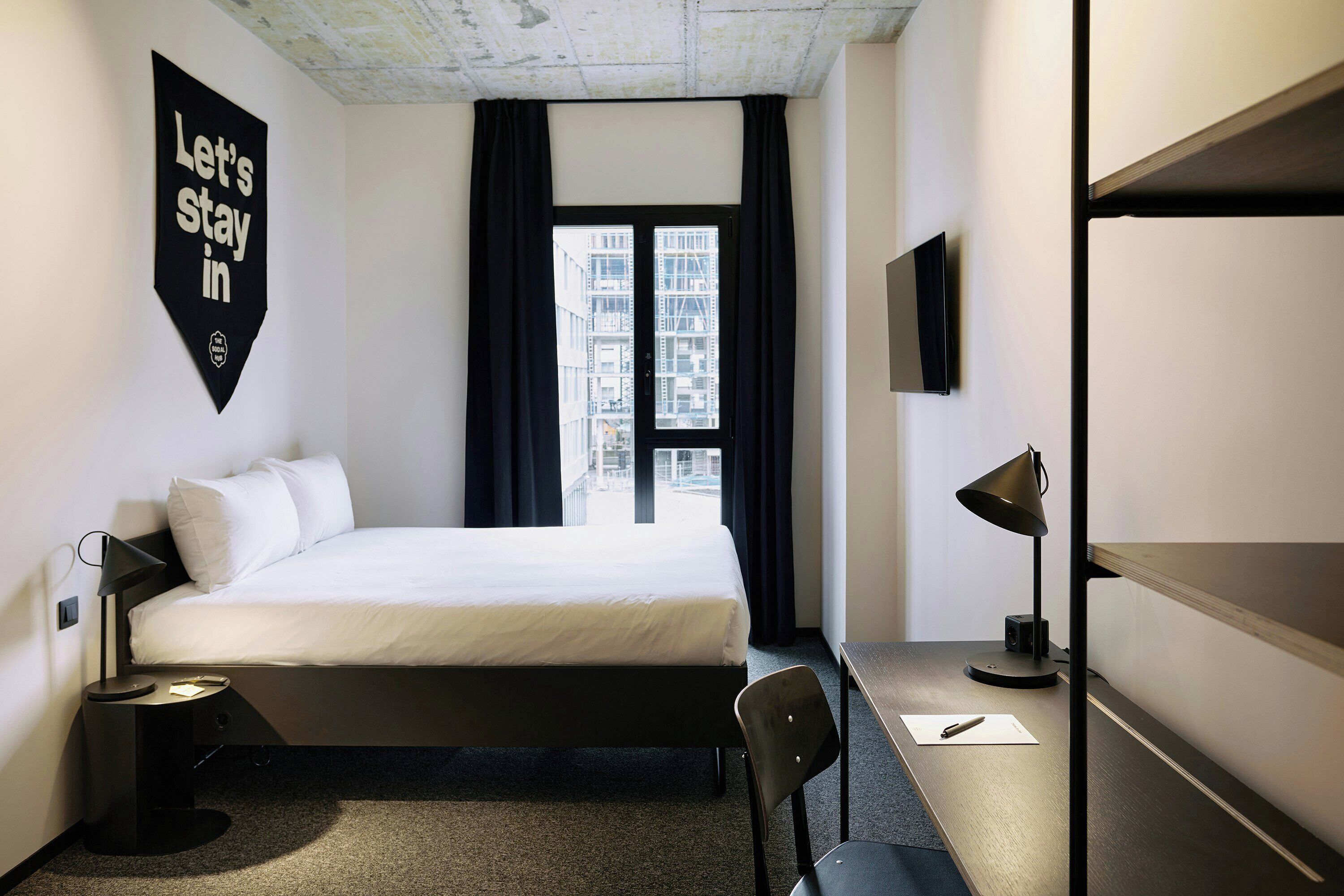 A hotel room with a bed, a desk, a chair, a desk lamp, a TV, and a window with curtains.
