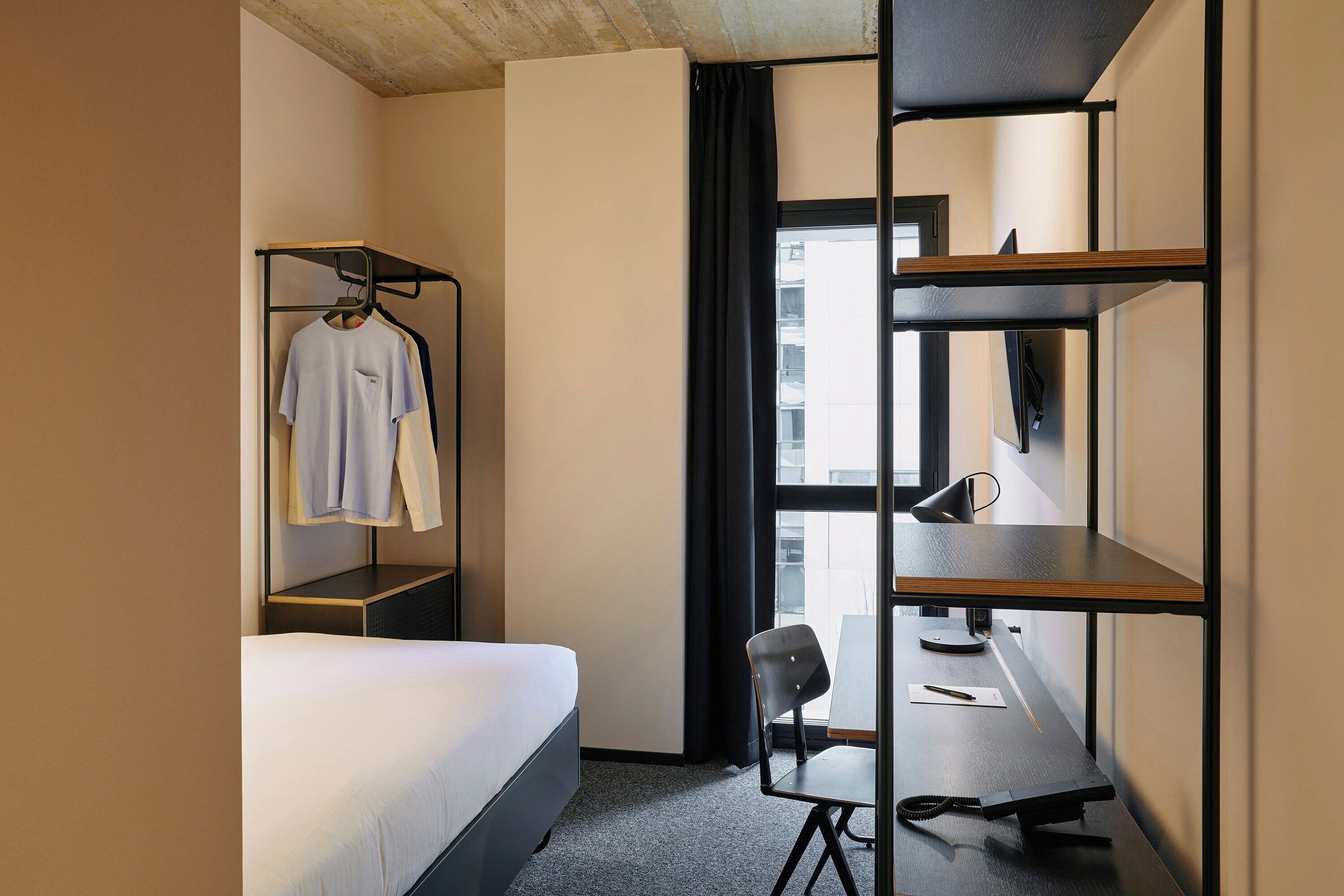 A hotel room with a bed, a desk with a chair, a coat rack, and a window with curtains.