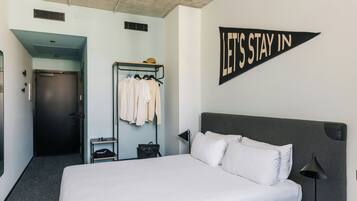 A hotel room with a bed, a wardrobe, a coat rack, a desk, and a 'LET'S STAY IN' sign.