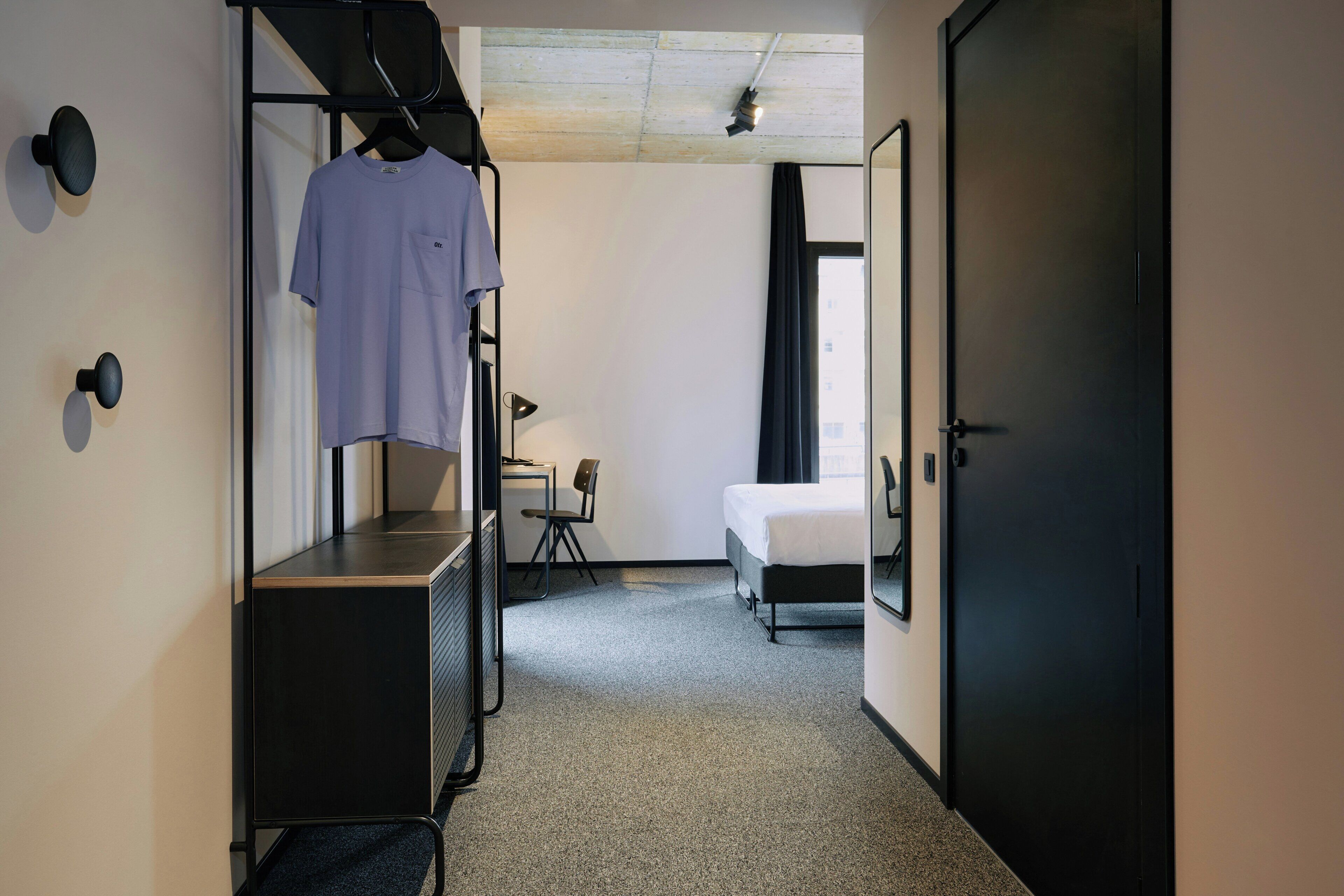 A hotel room with a bed, a desk with a chair, a wardrobe, and a hanging rack with a shirt.