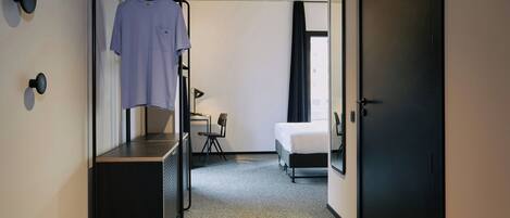 Et hotelværelse med en seng, et skrivebord med stol, et klædeskab og en bøjlestang med en skjorte.