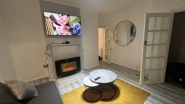 Ein Wohnzimmer mit Kamin, einem an der Wand befestigten Fernseher, einem runden Tisch mit Fernbedienung und einem Spiegel.
