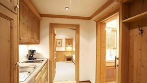 Un couloir étroit, avec des armoires en bois et un évier sur le côté gauche, mène à un espace cuisine équipé d’une cuisinière et d’autres armoires.