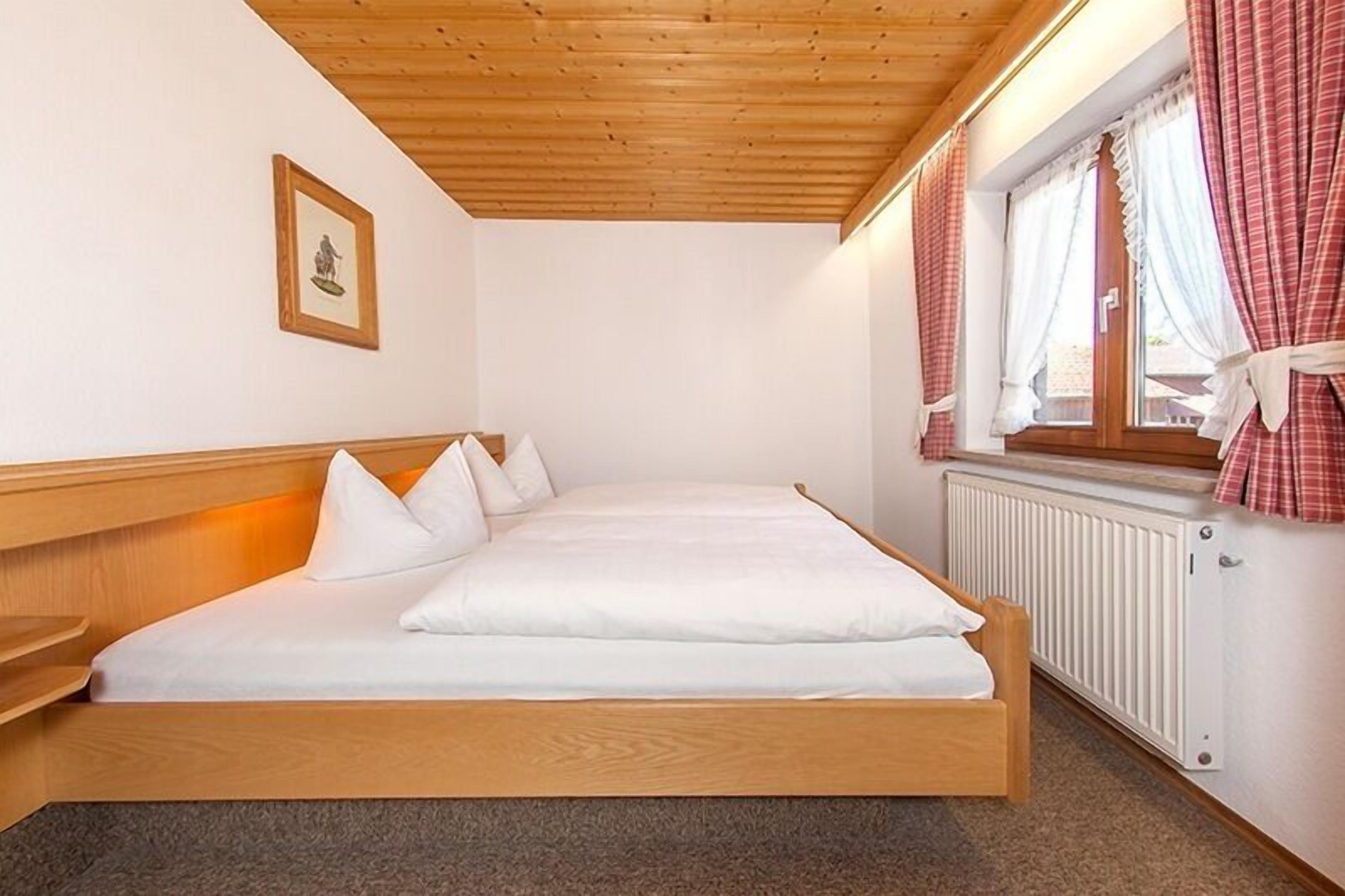 A single-bed room with a wooden headboard, a framed picture on the wall, and a window with red curtains.
