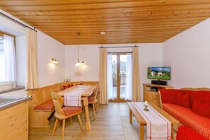 A cozy room with a wooden ceiling, a dining table with chairs, a sofa, a television, and a window with curtains.