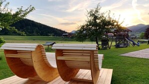 Deux bancs en bois, installés sur une plateforme en bois dans un parc, avec une aire de jeux et une vue sur une colline boisée.