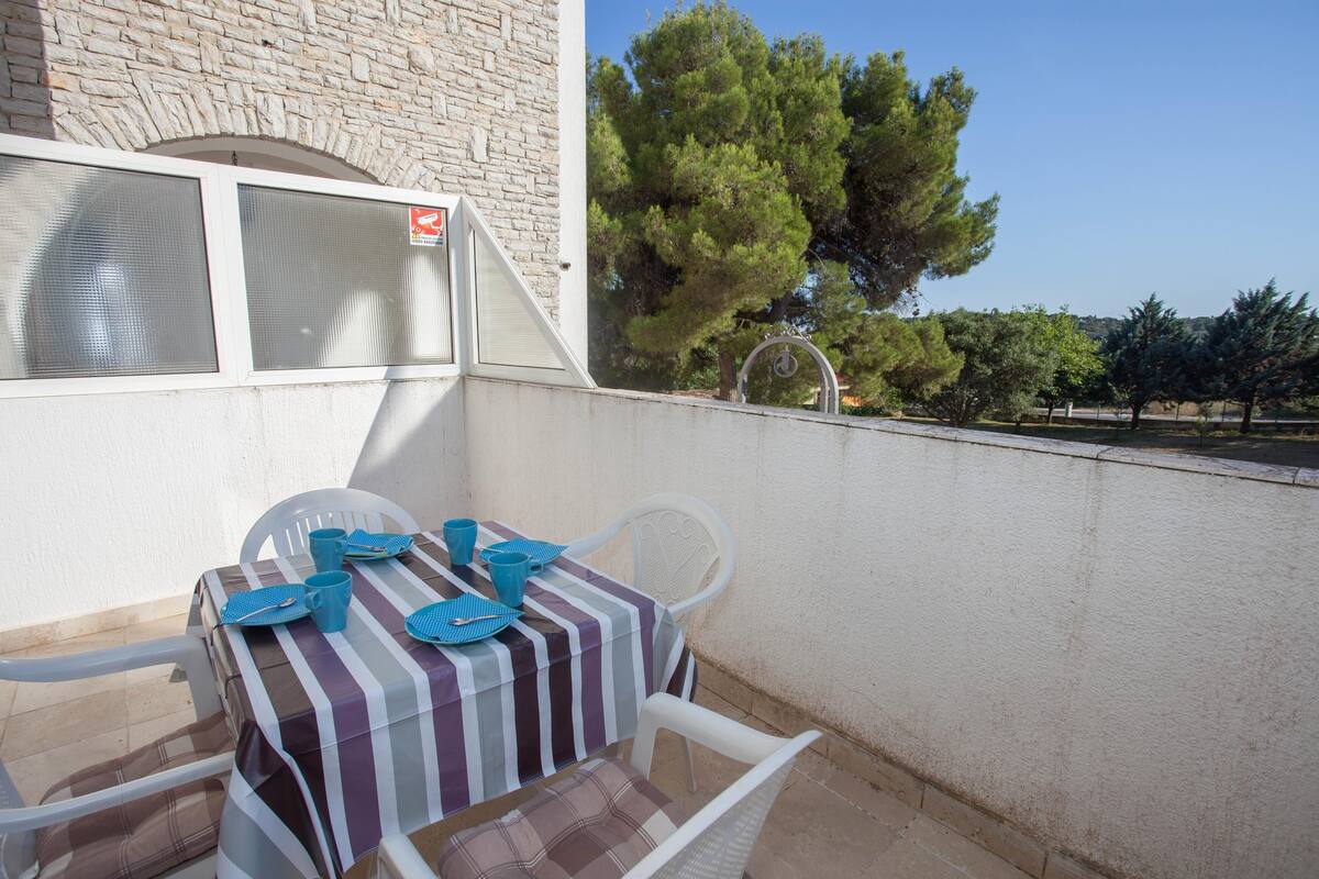 Un balcon avec une table dressée pour deux personnes, une chaise blanche et une vue sur des arbres et un immeuble.
