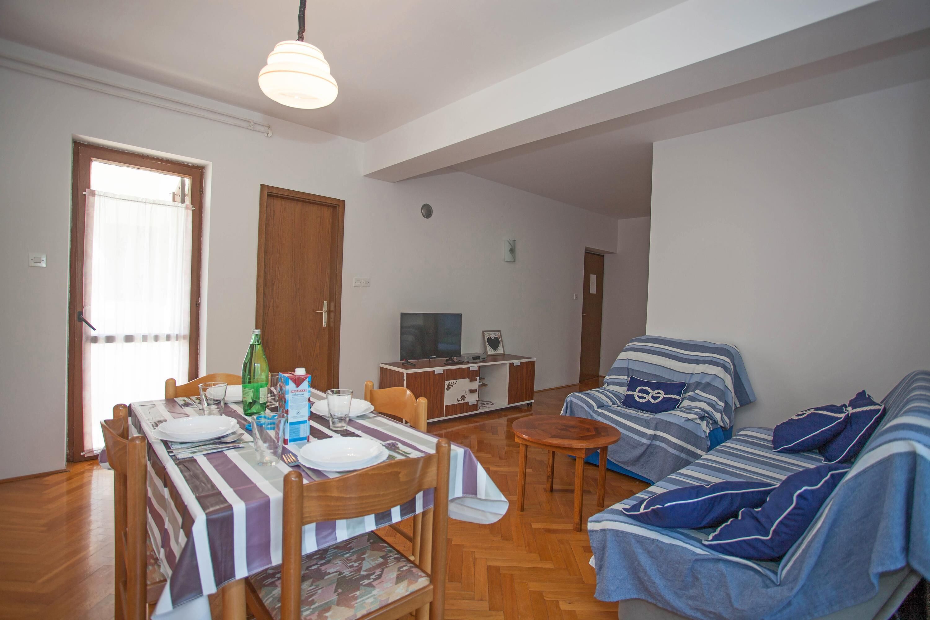 Uma sala de jantar com uma mesa posta para dois, um sofá e uma televisão ao fundo.