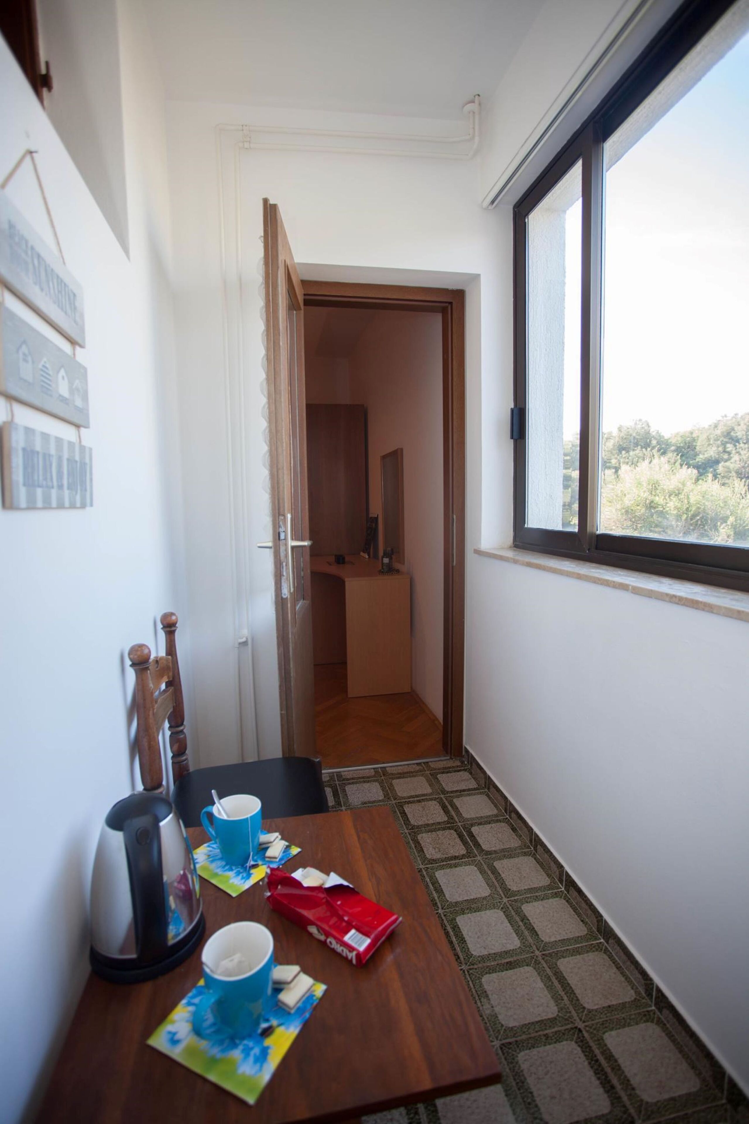 A narrow hallway with a wooden table set for two, a kettle, and a red box. A window on the right side offers a view of greenery outside.