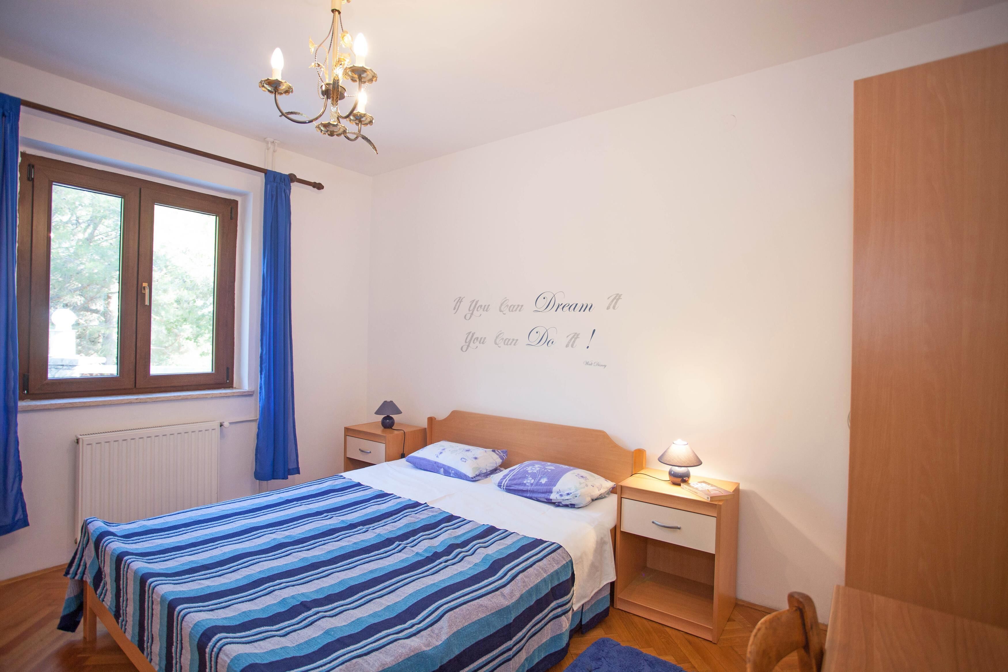 Un dormitorio con cama, mesitas de noche, candelabro, ventana con cortinas y una frase motivacional en la pared.