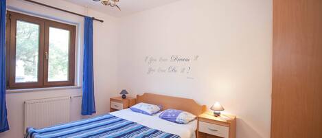 Un dormitorio con cama, mesitas de noche, candelabro, ventana con cortinas y una frase motivacional en la pared.