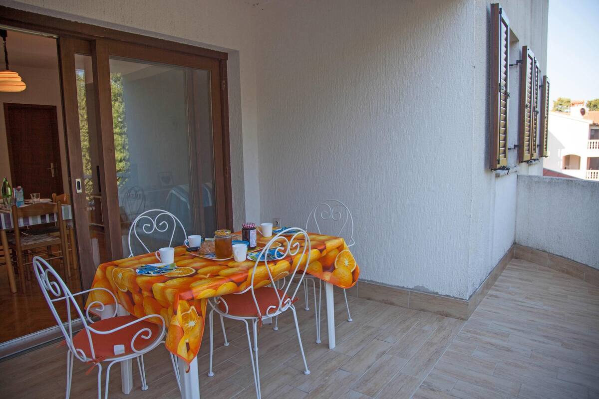 Un coin repas avec une table recouverte d’une nappe à motifs de fruits, des chaises en métal blanc et une porte-fenêtre coulissante donnant sur un espace extérieur.