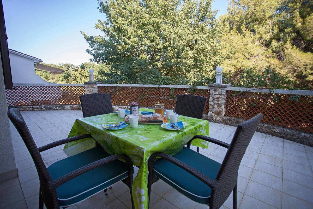 Une terrasse avec une table dressée pour deux personnes, entourée de chaises en osier agrémentées de coussins verts.