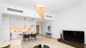 A modern living room with a large flat-screen TV, a dining area with a kitchen in the background, and a unique hanging light fixture. - Nasma Holiday Homes - Creek Horizon Tower 1 (Dubai)