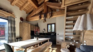 A rustic interior with a wooden dining table, bench, and sofa. A kitchen with a window and a staircase is visible.