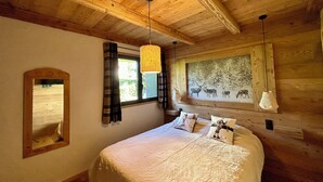 A wooden cabin bedroom with a bed, a window with curtains, a mirror, and a wall painting of deer.