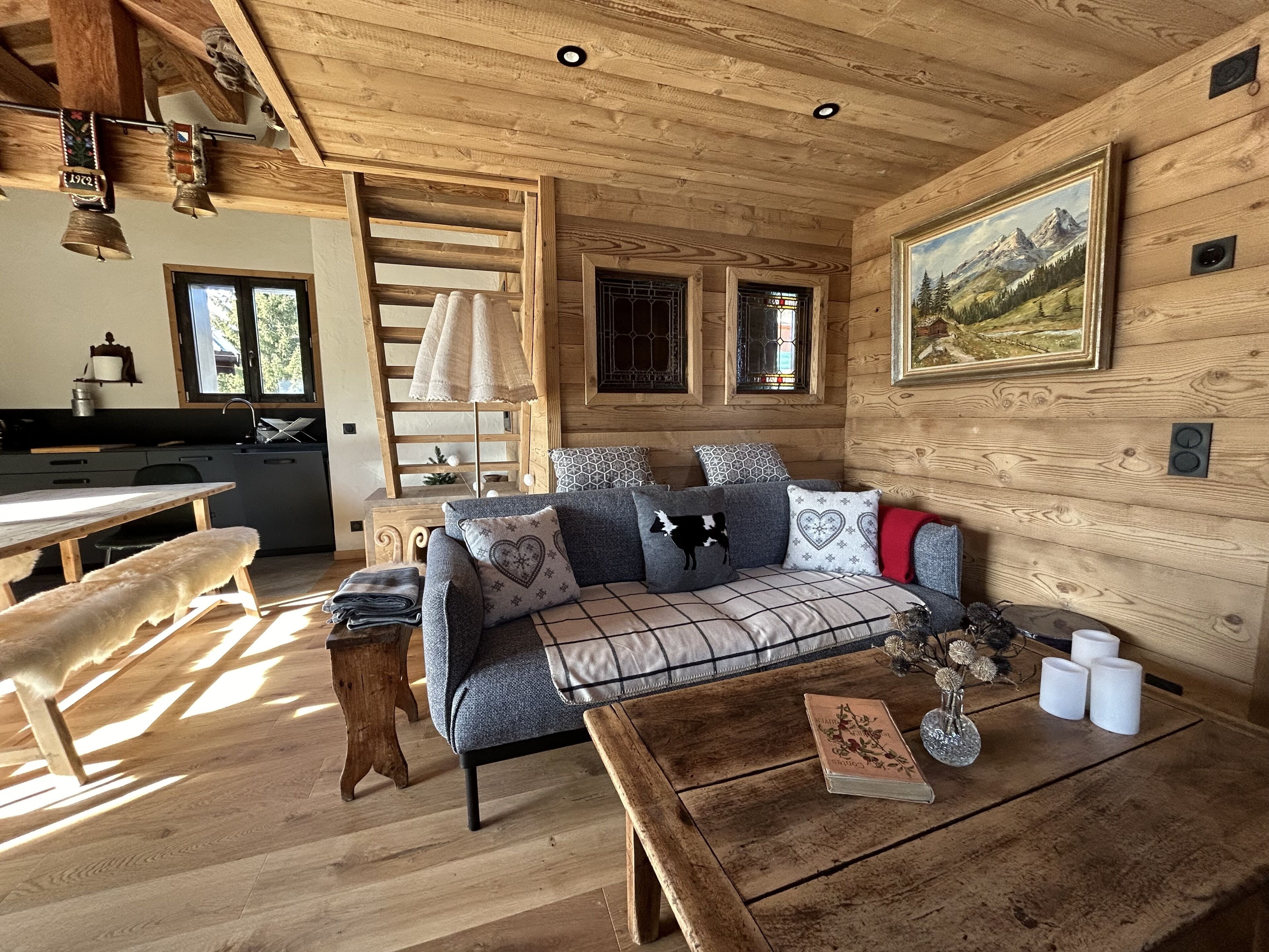 A cozy cabin interior with a wooden ceiling, a sofa with patterned cushions, a dining table, and a painting on the wall.
