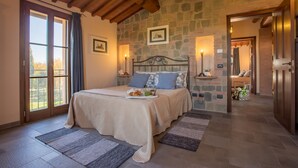 Une chambre avec un mur en pierre, un plafond en bois, un lit recouvert d’une couverture beige, une table de chevet avec une lampe, et une vue par la fenêtre.