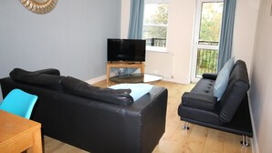 Ein Wohnzimmer mit schwarzem Sofa, einem Glastisch, einem Flachbildfernseher auf einem Schrank und einem Fenster mit Vorhängen.