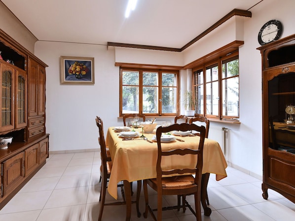 A dining room with a table set for four, wooden chairs, a clock on the wall, and a window with a view of trees.