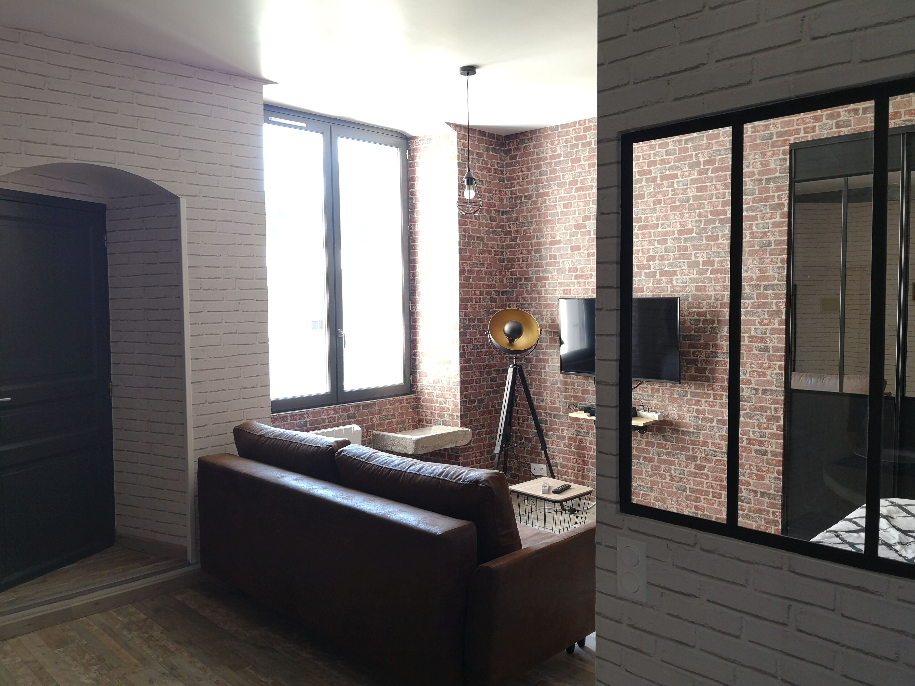 A modern living room with a brick wall, a large window, a sofa, a floor lamp, and a TV.