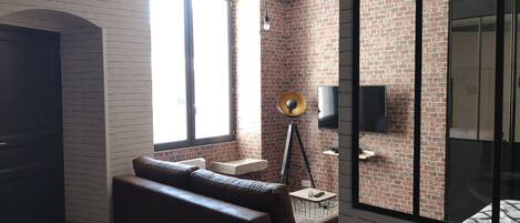 Una sala moderna con pared de ladrillo, un ventanal grande, un sofá, una lámpara de pie y un televisor.
