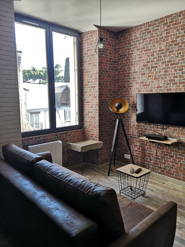 A living room with a brick wall, a large window, a leather sofa, a floor lamp, and a flat-screen TV.