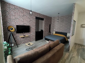 A room with a brick wall, a bed with a black headboard, a brown sofa, a small table with a lamp, and a TV mounted on the wall.