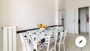 A dining area with a table covered by a colorful tablecloth, white chairs, and a bowl of fruit.