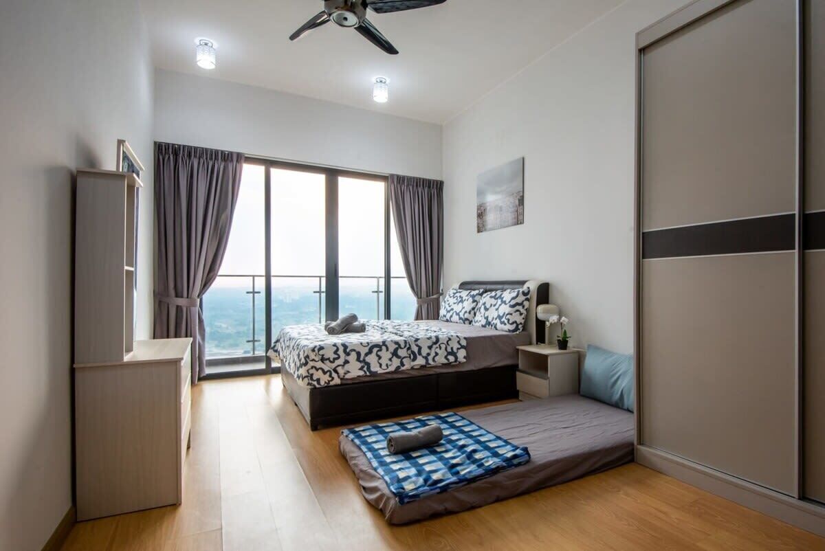 A modern bedroom with a large bed, a bedside table, a small desk, and a view of the outdoors through large windows.
