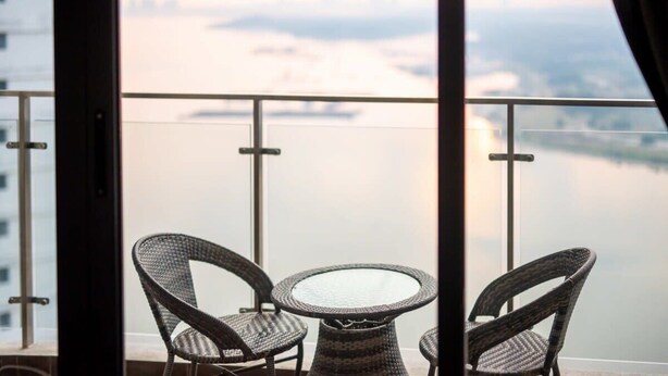 Ein Balkon mit Korbstühlen und einem Glastisch, mit Blick auf ruhiges Gewässer und eine ferne Stadtlandschaft.