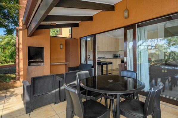 Een terras buiten met een ronde tafel en stoelen, een barbecue en uitzicht op de keuken.