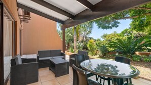 Un patio cubierto con mobiliario de mimbre y una mesa de vidrio, rodeado de abundante vegetación.