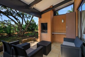 Een terras buiten met rieten meubilair, een ingebouwde barbecue en uitzicht op tropische begroeiing.