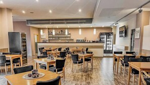 Um café moderno com mesas de madeira e cadeiras pretas, uma área de bar com prateleiras cheias de garrafas e um chão de madeira em padrão herringbone.