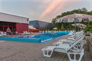 A swimming pool area with lounge chairs and umbrellas, a building with a red facade, and a blue roof. - Hotel Belaria Resort (Hradec nad Moravicí)