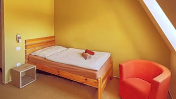 Una habitación pequeña con cama individual, techo inclinado y tragaluz, cabecera de madera, mesita de noche de madera y una silla roja.