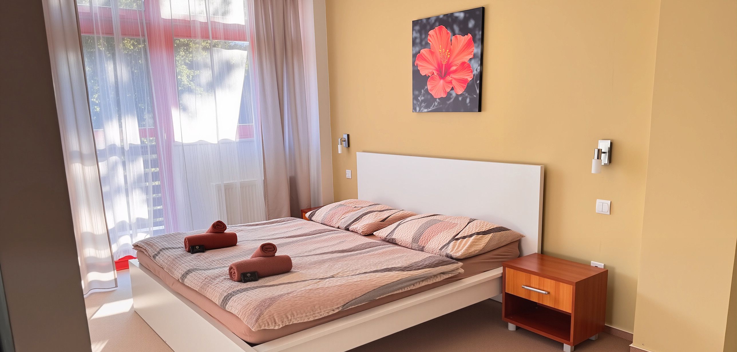 Una cama bien hecha con toallas enrolladas, una mesita de noche con lámpara y un cuadro de flores colgado en la pared.