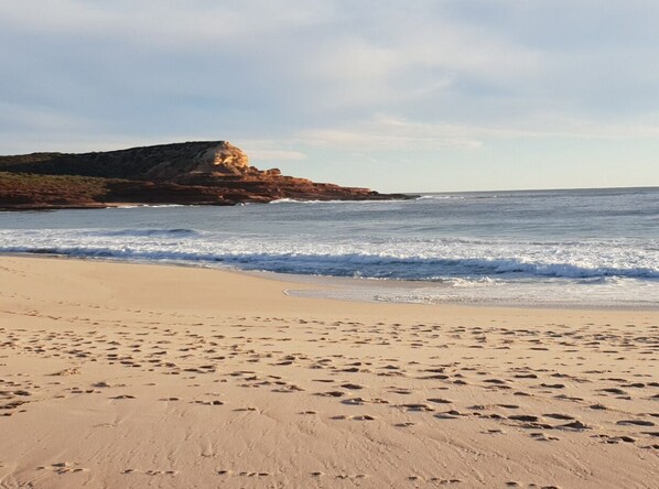 A sandy beach with footprints leading to the water, a rocky outcrop in the background, and a clear sky.