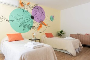 A hotel room with two beds, a wall mural, a small table with a lamp, and a wooden cabinet.