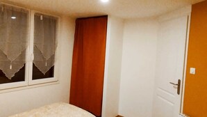 A small, simple room with a bed, a window with curtains, a door, and a light fixture on the ceiling. - Maisonnette à la Campagne " le Grain de Sel" (Hampont)