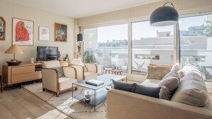 Ein modernes Wohnzimmer mit einer Couch, Couchtisch und Fernseher. An den Wänden hängen Bilderrahmen, und ein großes Fenster bietet Blick auf Gebäude.