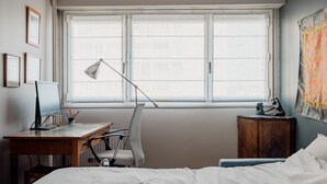 Ein Schlafzimmer mit einem Bett, einem Schreibtisch mit Computer, einem Stuhl, einer Lampe und einem Fenster mit Jalousien.