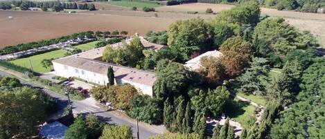 Vue aérienne d’un quartier résidentiel, avec une grande maison entourée d’arbres denses et de vastes champs.