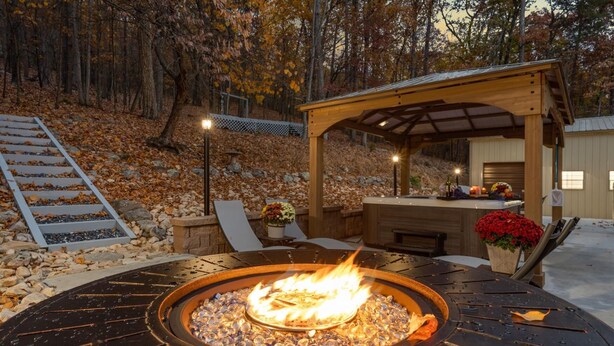 Un pozo de fuego circular al aire libre con una pérgola de madera y sillones de descanso al fondo.