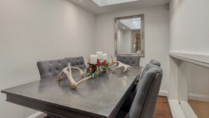 Une salle à manger avec une table haute, des chaises recouvertes d’un tissu gris, un arrangement floral en guise de centre de table et un grand miroir au mur.