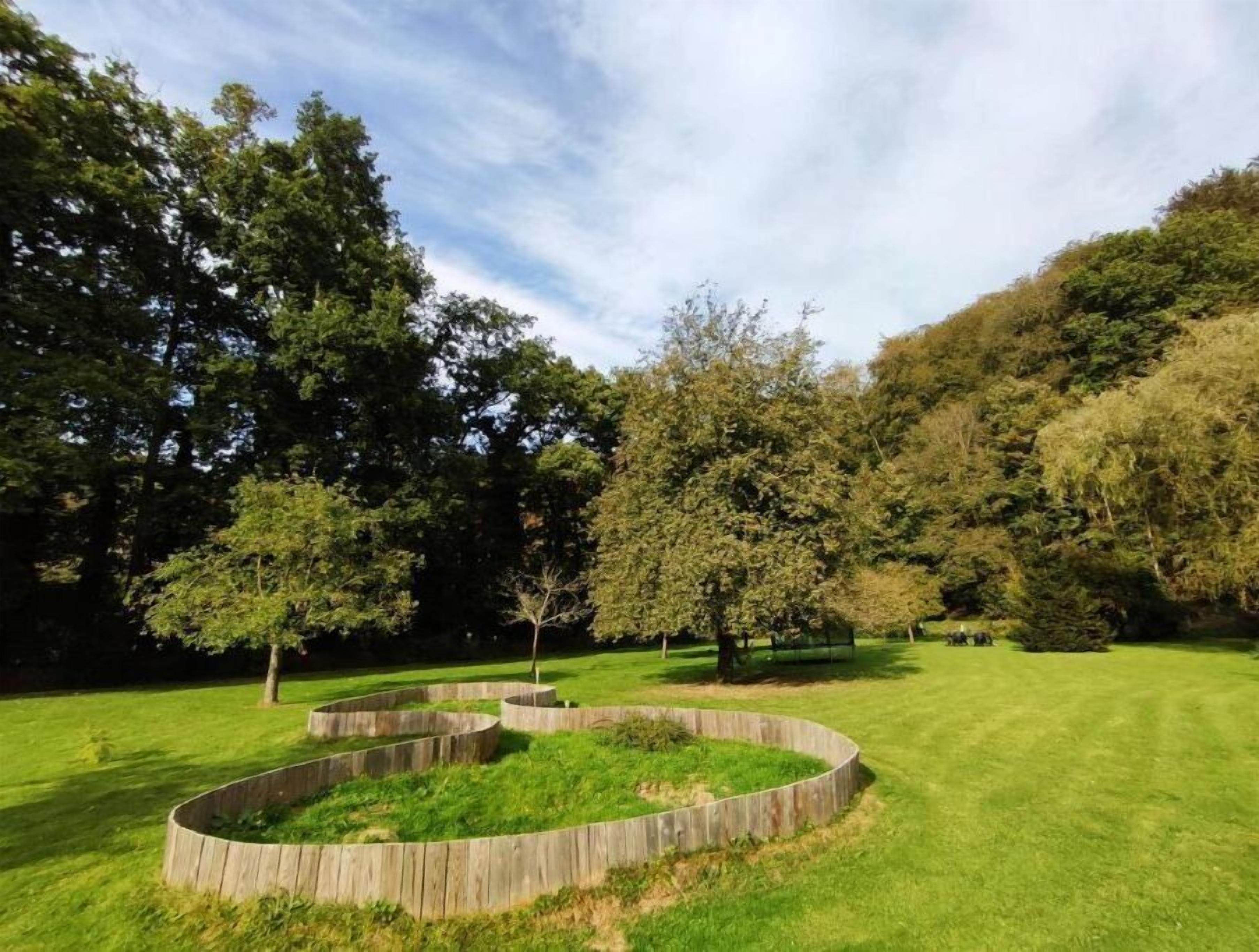 Een aangelegde omgeving met een ronde houten plantenbak, omringd door gras, bomen en een heuvel op de achtergrond.