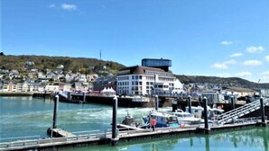 En hamn vid vattnet med förtöjda båtar, en modern byggnad med ett platt tak och en sluttning med hus i bakgrunden.