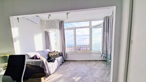 Een moderne kamer met een bank, een stoel, een klein tafeltje en een groot raam met uitzicht op de lucht en gebouwen.
